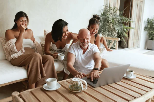 Família feliz com laptop se divertindo juntos. Pai, mãe e filhas sentados no sofá no pátio, bebendo chá, usando computador portátil e desfrutando de lazer no resort tropical. — Fotografia de Stock