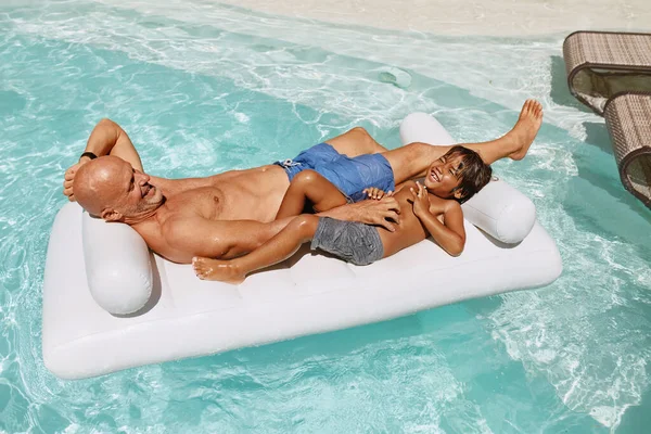 Apa és fia napoznak a medencében. Kisfiú apával a felfújható matracon. A szülők és a gyerekek élvezik a nyaralást az üdülőben. Családi hétvége Villában. Nyári szabadidő mint életstílus. — Stock Fotó