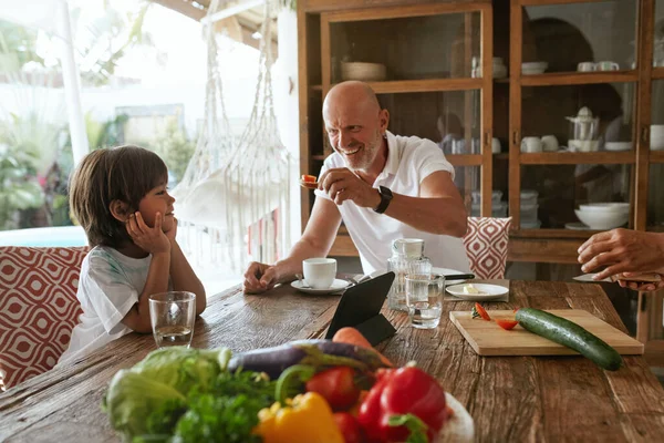 Padre e hijo comiendo en la mesa de la cocina. Padres y niños comiendo comida vegetariana en Villa en Tropical Resort. Familia positiva disfrutando de las vacaciones de verano. Dieta saludable como estilo de vida. — Foto de Stock