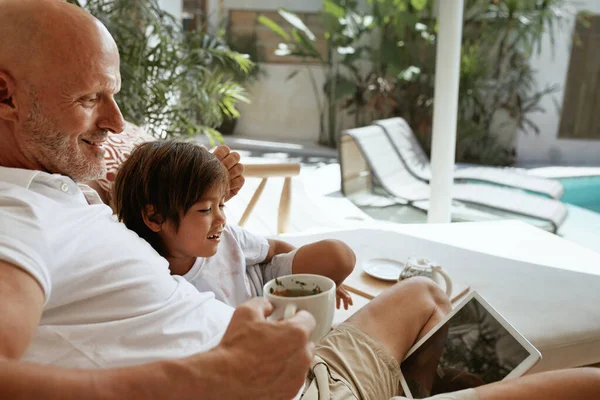 Acara keluarga di rumah. Dad And Little Boy Sitting On Sofa In Tropical Villa Patio With Tablet (dalam bahasa Inggris). Ayah Meminum Teh dan Menggunakan Perangkat Digital Portabel. Nikmati Ieisure On Weekend at Resort. — Stok Foto