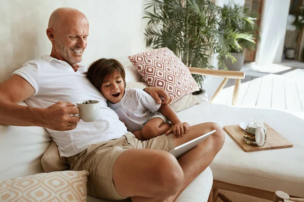 Baba ve Oğul, Villa 'da aile hafta sonlarında tablet kullanıyor. Mutlu baba ve küçük çocuk koltukta oturuyor, taşınabilir dijital cihaz kullanıyor ve evde eğleniyor. Tropikal Tatilde Yaz Tatili. — Stok fotoğraf