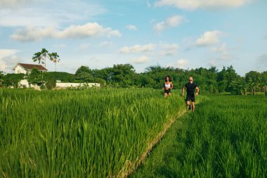 Sabah pirinç tarlasında koşan olgun bir çift. Beyaz adam ve Asyalı kadın tropikal manzaraya karşı yeşil çimlerin arasında koşuya çıktılar..