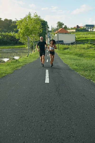 Pareja madura corriendo en el camino en el paisaje tropical. Caucásico hombre y asiático mujer en trotar entrenamiento en la mañana cerca de verde arroz campos. —  Fotos de Stock
