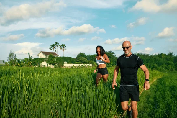 Pareja madura corriendo en el campo de arroz por la mañana. Caucásico hombre y asiático mujer en trotar entrenamiento entre verde hierba contra tropical paisaje. —  Fotos de Stock
