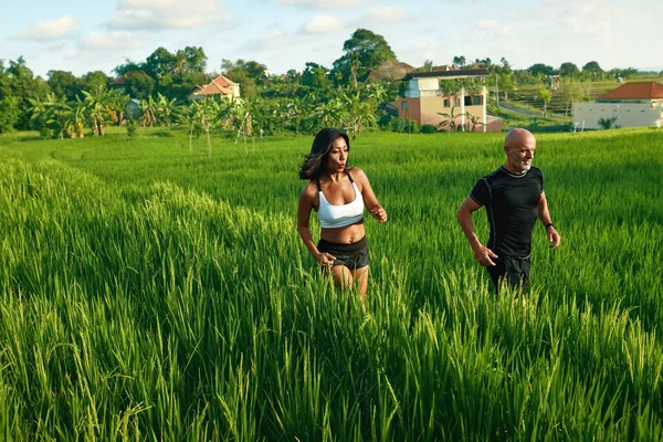 Casal maduro correndo no campo de arroz de manhã. Homem caucasiano e mulher asiática em jogging treino entre grama verde contra paisagem tropical. — Fotografia de Stock