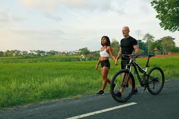 Deportiva pareja caminando bicicleta en la carretera. Mujer asiática y hombre caucásico caminando por las manos con bicicletas y hablando en contra de verde paisaje tropical. —  Fotos de Stock