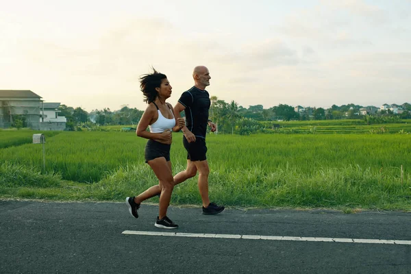 Couple d'âge mûr courant sur la route au paysage tropical. homme caucasien et asiatique femme sur jogging séance d'entraînement dans le matin près vert riz champs. — Photo