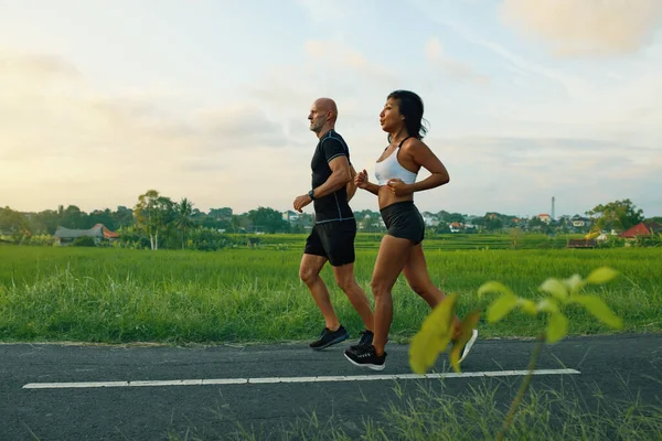 Pareja madura corriendo en el camino en el paisaje tropical. Caucásico hombre y asiático mujer en trotar entrenamiento en la mañana cerca de verde arroz campos. —  Fotos de Stock