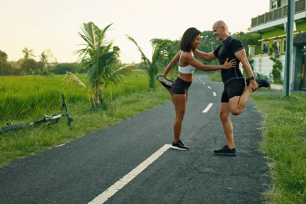 Un par de calentamiento antes de correr por la carretera. asiático mujer y caucásico hombre haciendo pareja estiramiento antes de trotar en naturaleza. —  Fotos de Stock