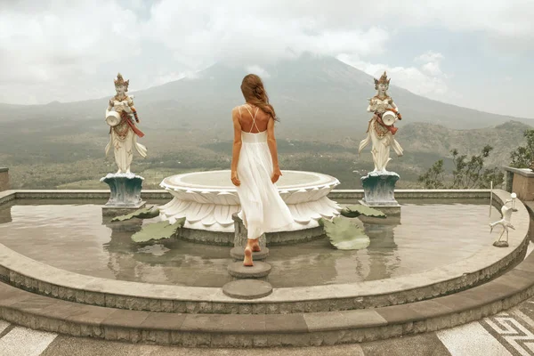 Beautiful Girl Posing On Fountain Against Agung Volcano In Bali, Indonesia. Young Woman In White Dress Fluttering In Wind Looking At Mountain Peak In Clouds From Terrace. — Stock Photo, Image