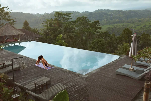 Menina bonita fazendo Yoga na beira da piscina de manhã em Bali, Indonésia. Jovem Mulher Magro em Sportswear em pé em virada para baixo Dog Pose no tapete desportivo Perto Infinito Piscina Contra a paisagem tropical. — Fotografia de Stock