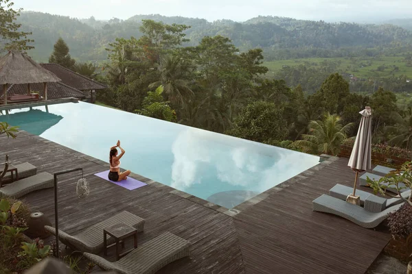 Menina bonita fazendo Yoga na beira da piscina de manhã em Bali, Indonésia. Mulher magra nova no Sportswear que senta-se na dose do lótus ou Padmasana no tapete do esporte perto da associação do infinito de encontro à paisagem tropical. — Fotografia de Stock