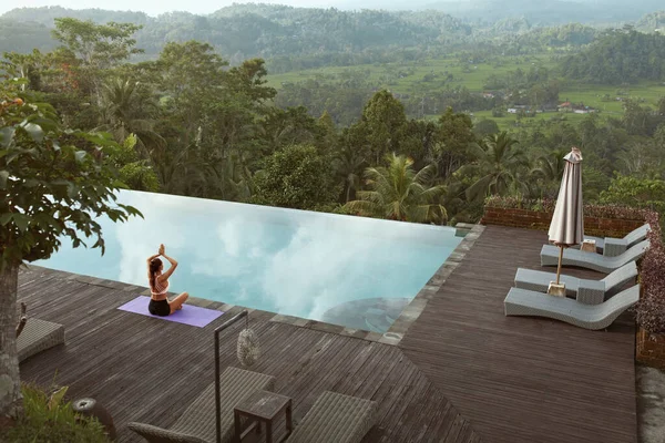 Menina bonita fazendo Yoga na beira da piscina de manhã em Bali, Indonésia. Mulher magra nova no Sportswear que senta-se na dose do lótus ou Padmasana no tapete do esporte perto da associação do infinito de encontro à paisagem tropical. — Fotografia de Stock