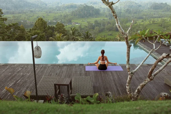 Hermosa chica haciendo yoga en la piscina por la mañana en Bali, Indonesia. Joven mujer delgada en ropa deportiva sentada en loto Pose o Padmasana en la alfombra deportiva cerca de la piscina infinita contra el paisaje tropical. — Foto de Stock