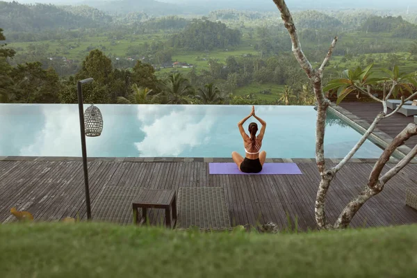 Menina bonita fazendo Yoga na beira da piscina de manhã em Bali, Indonésia. Mulher magra nova no Sportswear que senta-se na dose do lótus ou Padmasana no tapete do esporte perto da associação do infinito de encontro à paisagem tropical. — Fotografia de Stock