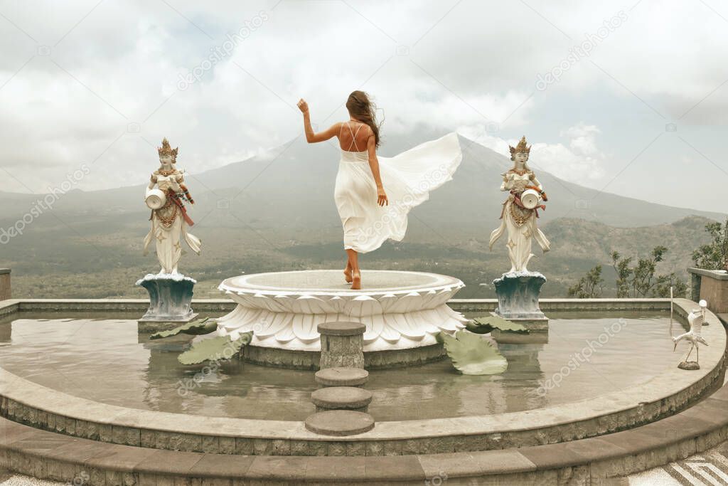 Beautiful Girl In White Dress Fluttering In Wind Looking At Agung Volcano In Bali, Indonesia. Young Woman Walking Barefoot On Fountain And Posing Between Sculptures.