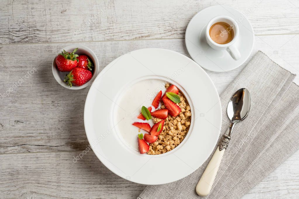 close-up photo of oatmeal with fresh strawberries and green mint leaves with milk in white plate on grey wooden table with cup of coffee