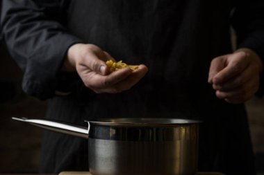 Bir şef makarna penne bir avuç su, ahşap kesme tahtası koyu bir arka plan ev yemekleri kavramı ile metal bir tava üzerinde tutan eller Close-up