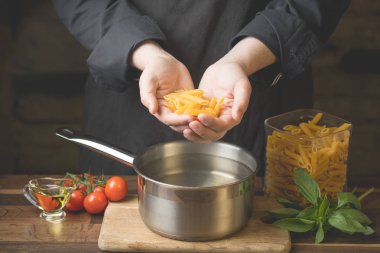 Bir şef makarna penne bir avuç su, ahşap kesme tahtası koyu bir arka plan ev yemekleri kavramı ile metal bir tava üzerinde tutan eller Close-up