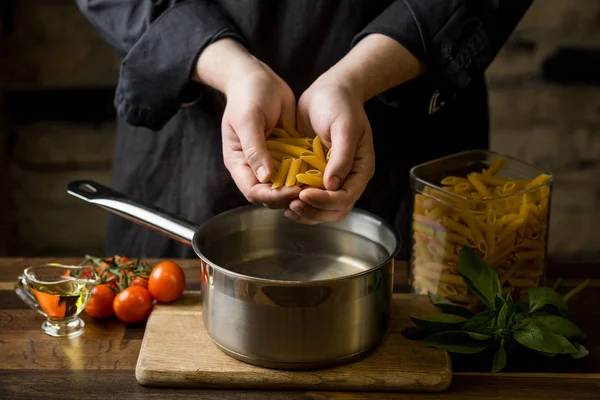 Bir şef makarna penne bir avuç su, ahşap kesme tahtası koyu bir arka plan ev yemekleri kavramı ile metal bir tava üzerinde tutan eller Close-up