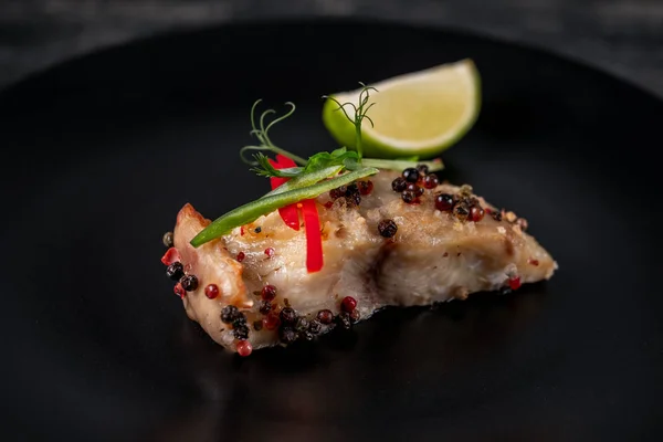 Close view of fresh cooked fish slice baked in pepper grains served with lime slice and herbs on black plate