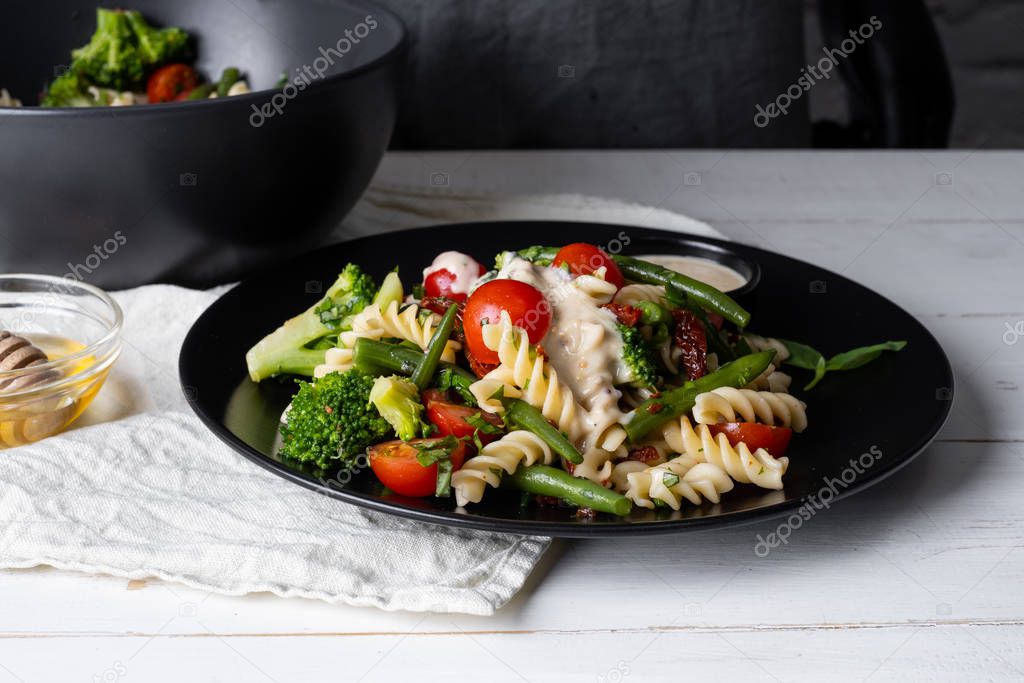 Bowl and plate full of warm salad with pasta and vegetables on white wooden table