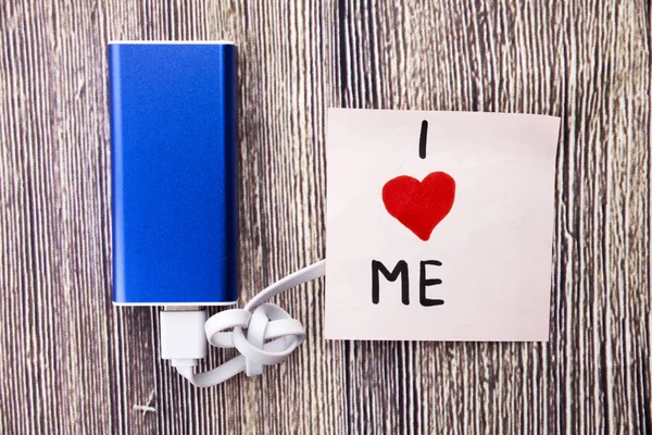 I love me heart handwritten on note paper in different colors and heart shape in red color. Energy storage device of blue color with cable on wooden background. selfish or self love Concept.