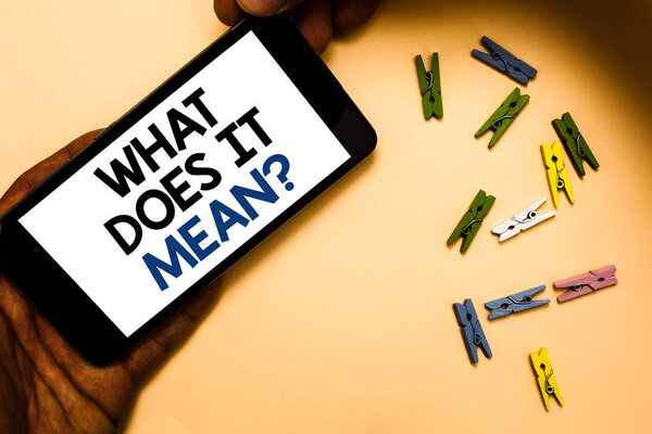 Conceptual hand writing showing What Does It Mean question. Business photo text Give me the meaning of something Definition Hand holding iPhone with letters and paperclip peach color ground
