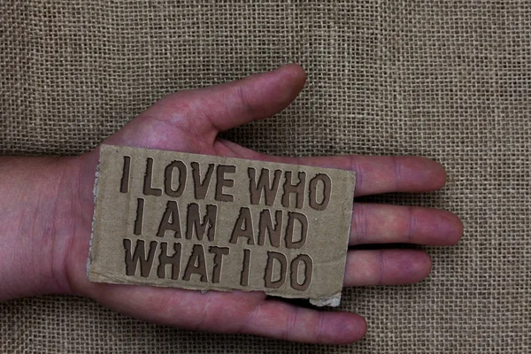 Conceptual hand writing showing I Love Who I Am And What I Do. Business photo text High self-stem being comfortable with your job Cardboard with marked some letters on human hand jute sack ground. — Stock Photo, Image