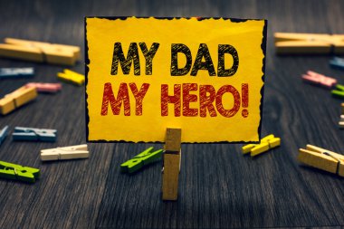 Word writing text My Dad My Hero. Business concept for Admiration for your father love feelings emotions compliment Blacky wooden desk laid paper clip randomly one hold yellow board with text.