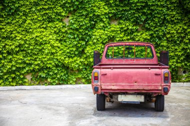 Rustic Red Pickup Parked Alone in Open Lot. Abandoned Old Truck with Stake Rail Board Facing a Shrubbery Wall. Open Delivery Vehicle Shot at Rear End View. clipart
