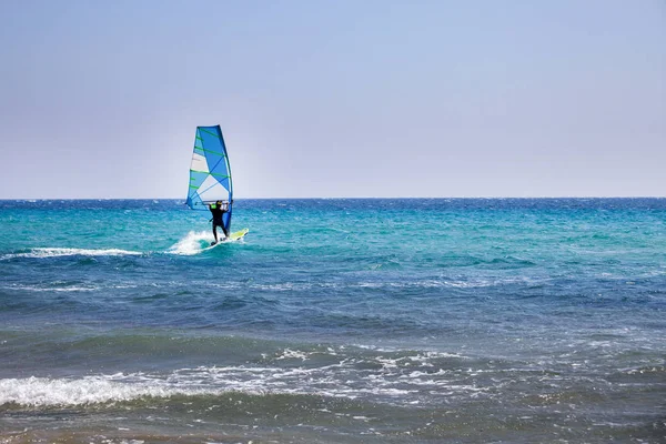Surfer Spending Time on Outdoor Water Sport Adventure. Clear Sky and Blue Wave. Person Balancing on a Windsurfing Board with Sail. Vacation Activity ideas.