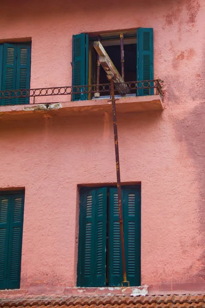 Rustic Iron Steel Supporting a Wood Plank Hanging Horizontally from High Windowsill. Old Apartment Building with Pink Wall and Blue Shutter Needing Repair.