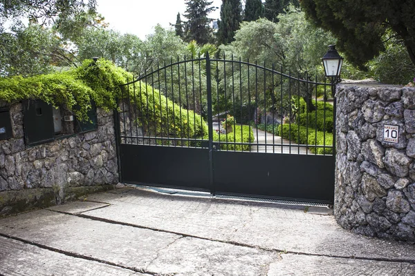 High Iron Gated Driveway Entrance of a Villa. Architectural Garden Landscape Surrounding the Curbed Pathway. Stone and Rock Paved Wall Securing the Estate.