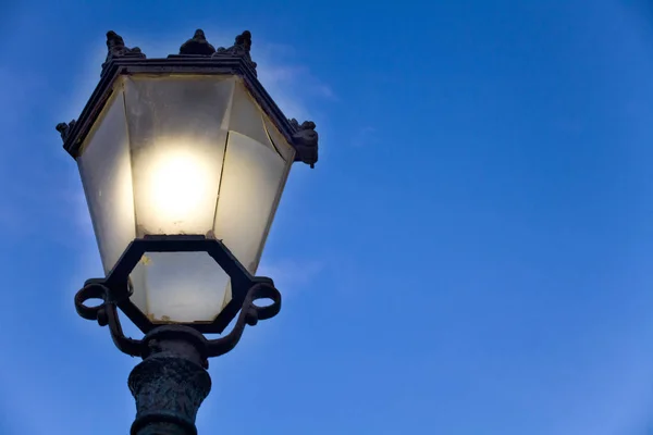 Poste de lámpara de estilo victoriano con bombilla iluminada y panel de vidrio roto. Linterna de calle al aire libre hecha de fundición de hierro forjado negro bajo un cielo azul . —  Fotos de Stock