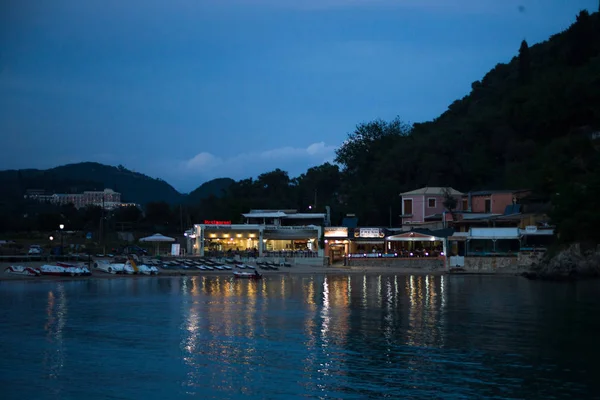 Water Reflecting Lights From Restaurants. Eateries are Operating at Dusk time. Pedal Boats, Recliners on Sand Facing the Ocean. Verdant Hills at the back.