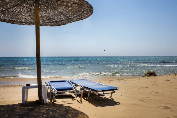 Paraguas de madera Proyección de la luz del sol y el calor. Reclinador de playa plegable y desplegable ajustable frente al océano. Arena fina y agua azul en un día soleado . — Foto de Stock