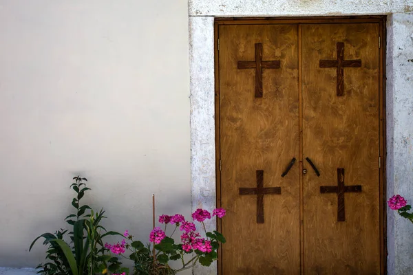 Four Crosses Mounted on Two Panel Closed Door. All Materials are made of Wood against White Cemented Wall. Plants with Pink Flowers Placed at the Entrance.