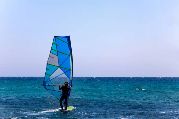 Jezdec v neoprénový oblek a helmu vyvážení na surfovací prkno s plachtou stožár a Bar. Jasná obloha a modré vlny. Surfař vyzkoušet Outdoor Adventure Sport vodní. — Stock fotografie