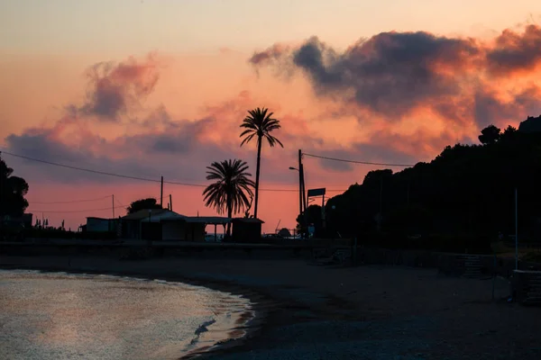 Céu rosa e cinza que olha sobre o litoral da praia. Noite a começar a cair. Casas e árvores Criando uma forma de silhueta. Água refletindo a cor do crepúsculo . — Fotografia de Stock