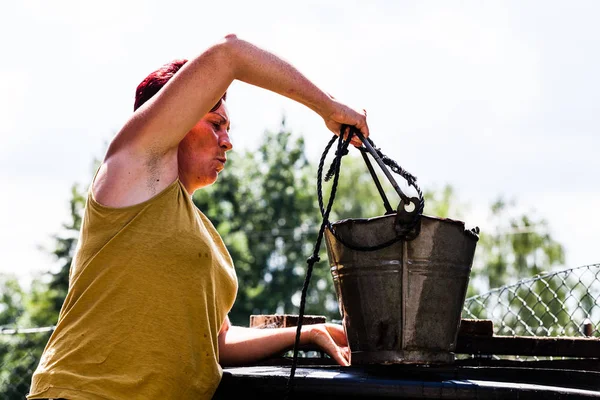 Boční pohled ženy držící plechový kbelík s řetězcem. Lady pracovní tvrdé načítání z přírodních studní. Typická denní práce v domácnosti. Scénář živé přírody. — Stock fotografie
