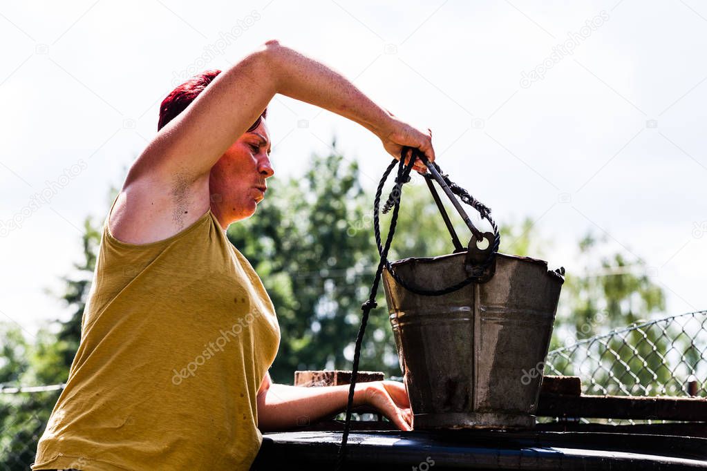 Side view of a woman holding a metal bucket with a string. The lady working hard fetching water from the natural well. Typical daily household chore. Countryside living scenario.