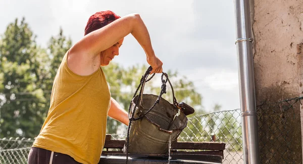 Una mujer que trabaja duro buscando agua del pozo natural. La madre de mediana edad tirando del recipiente de metal con una cuerda. Típica tarea diaria del hogar. Escenario de vida del campo . —  Fotos de Stock