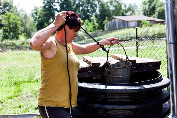 Vista lateral de una madre trabajadora sosteniendo un cubo de metal. La mujer apuntando el contenedor al centro del pozo natural. Buscando agua potable para beber. Concepto de vida rural . —  Fotos de Stock