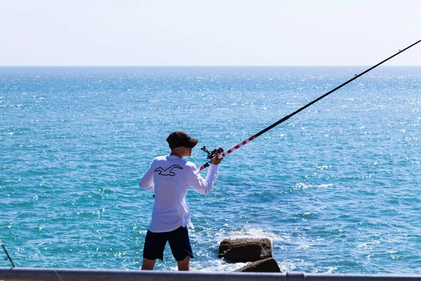 Брайтон, Великобритания - июнь 2018 года Человек с капюшоном, стоящий на берегу и держащий длинную удочку с катушкой. Кристально-голубая волнистая вода в солнечный день. Идеи для занятий водными видами спорта и отдыха . — стоковое фото