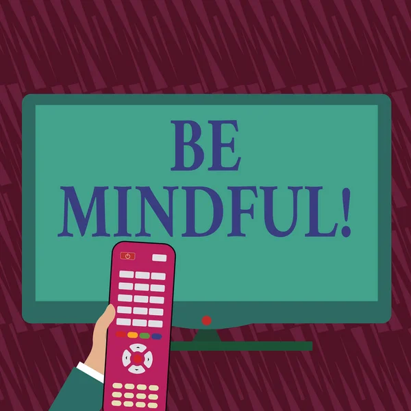 Text sign showing Be Mindful. Conceptual photo Asking demonstrating become conscious or aware of something Hand Holding Computer Remote Control infront of Blank Wide Color PC Screen.