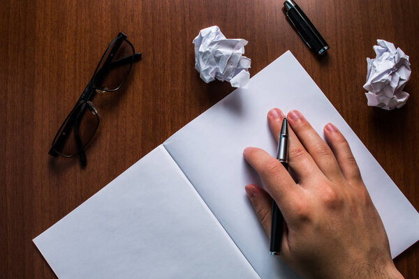 Upper close view man hand resting pen open notebook reading glasses lying wooden table timber desktop. Writing down essay idea project goal homework note