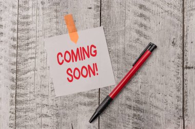 Text sign showing Coming Soon. Conceptual photo event or action that will happen after really short time One plain blank paper sheet and a pen attached with wooden textured table.