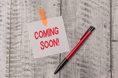 Text sign showing Coming Soon. Conceptual photo event or action that will happen after really short time One plain blank paper sheet and a pen attached with wooden textured table. clipart