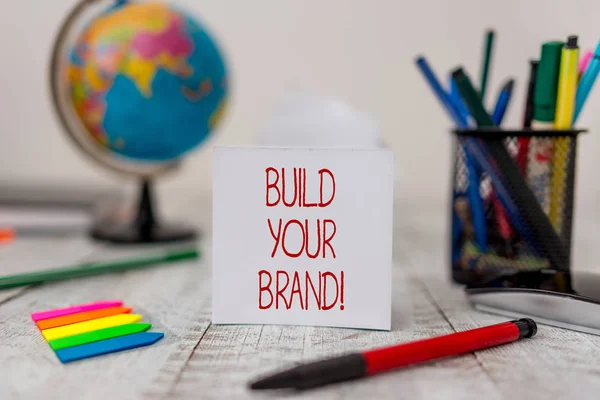 Sinal de texto mostrando Construir sua marca. Foto conceitual cria ou melhora o conhecimento dos clientes e opiniões do produto Folhas estacionárias e de papel, além de pequenas setas com globo na mesa de madeira . — Fotografia de Stock